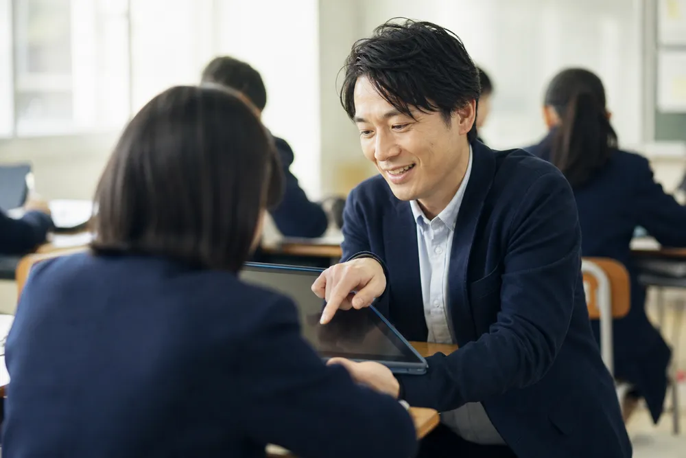 児童の隣にかがみ、タブレットの画面を指しながら教える先生（イメージ）